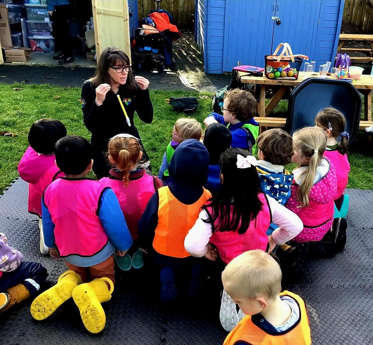 A lady is telling a story to a group of nursery children outside in a garden.
