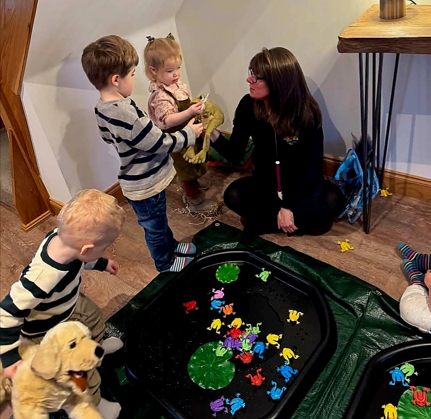A lady is holding a frog puppet and interacting with two toddlers. There are toy frogs in a black tuff tray.