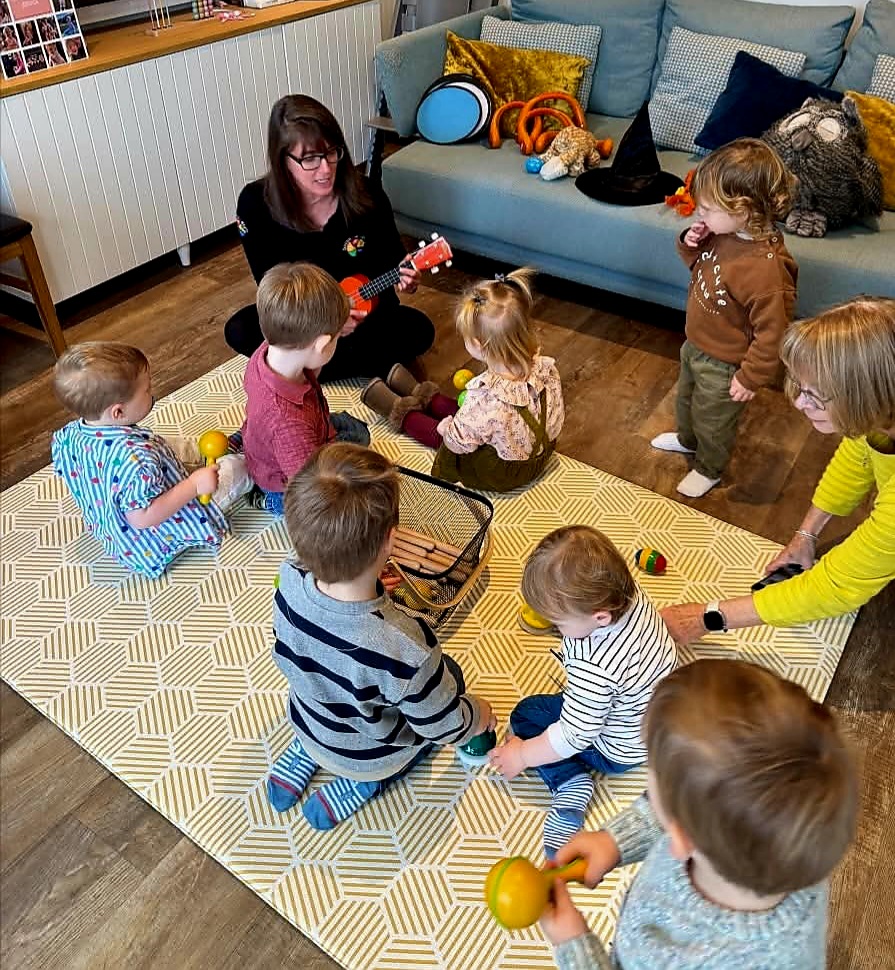 A lady plays the ukulele while children play along with maracas and bells