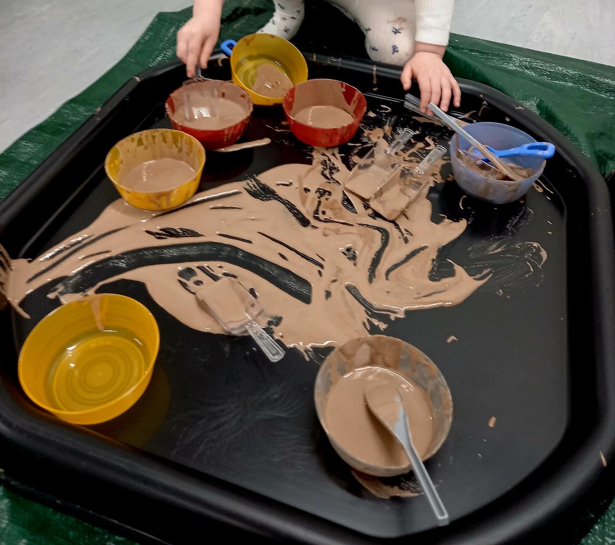 A child's busy hands are playing with bowls, spoons and chocolate mousse in a black tuff tray.