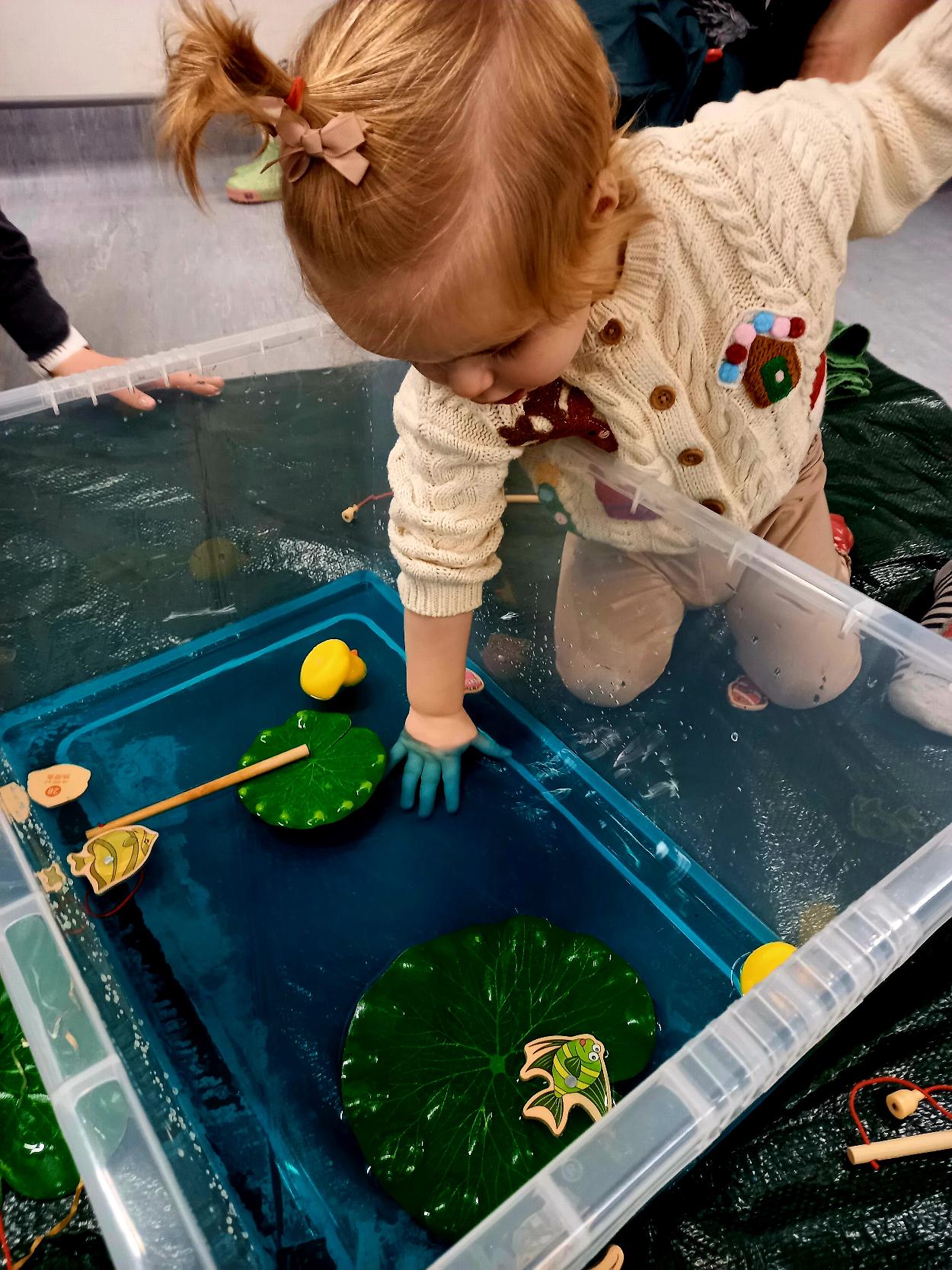 A toddler plays with coloured water, fish and ducks in a big basin.